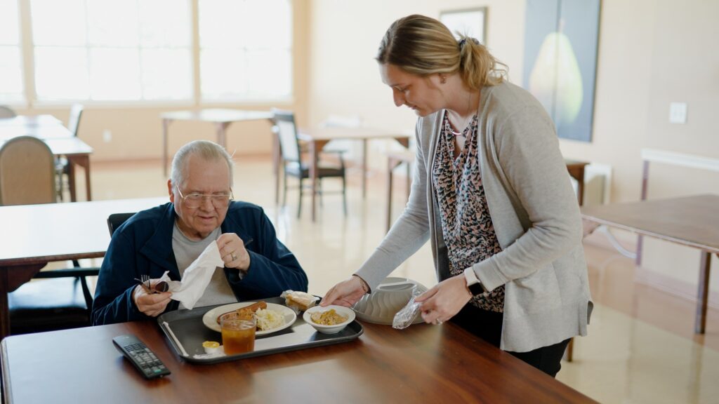 A long-term care resident is served dinner by a kitchen staff worker at a skilled nursing facility