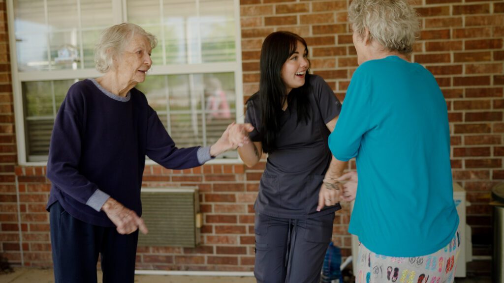 two women and a nurse smile and hold hands at a skilled nursing facility