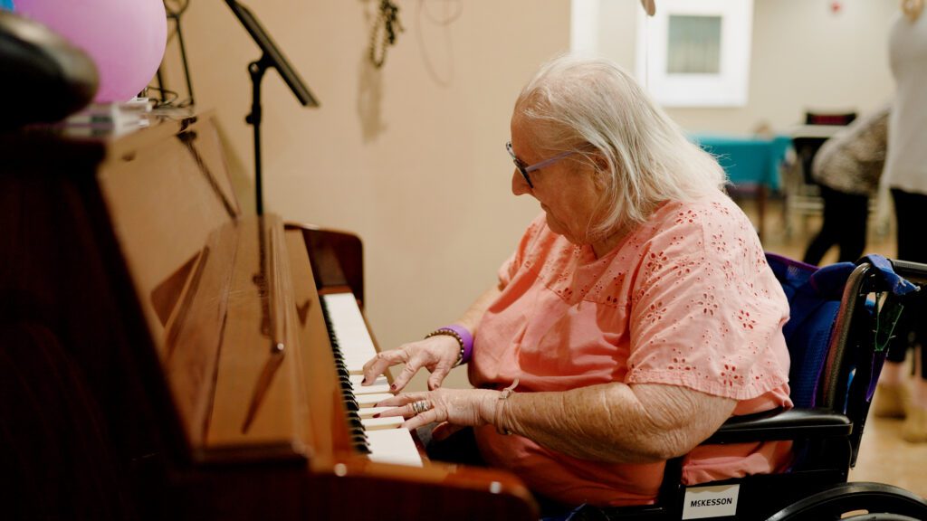 A woman plays the piano at a skilled nursing facility during a respite care visit